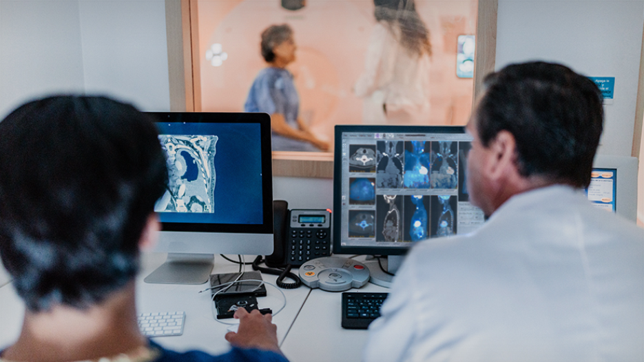 Image of two clinicians looking at radiography images on a computer screen with a patient and another clinician visible in a room behind them.